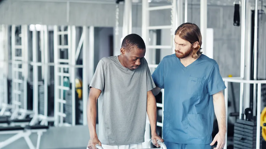 A man relearning to walk with a wheeled walker while a therapist steadies him