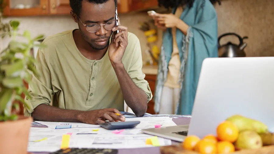 A man on the phone at his kitchen table with a calculator and paperwork in front of him