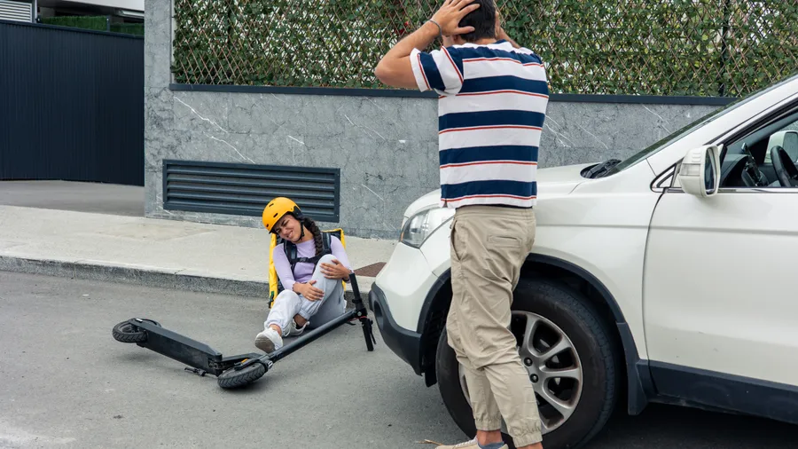 A delivery courier on the ground beside a toppled scooter while the other driver stands by his car